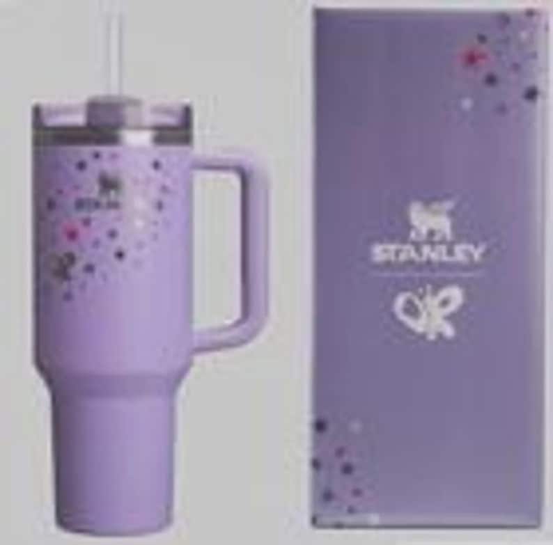 K&ouml;nnte beinhalten: Ein lavendelfarbener Stanley-Becher mit Griff, verziert mit bunten Punkten und einem Schmetterlingsdesign. Der Becher hat einen Deckel und einen Strohhalm. Eine passende lavendelfarbene Box mit dem Stanley-Logo und Schmetterlingsdesign befindet sich neben dem Becher.