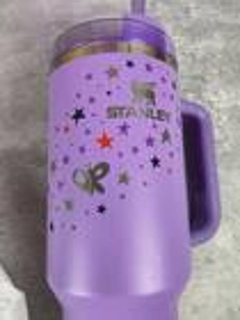 Puede incluir: Un vaso Stanley morado con asa y tapa transparente. El vaso presenta estrellas doradas y el logotipo de Stanley. El dise&ntilde;o incluye una mariposa dorada y el texto "OR".