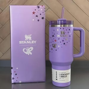 Puede incluir: Un vaso Stanley Quencher H2.0 Flowstate lavanda con asa y pajita, junto a una caja a juego. El vaso y la caja est&aacute;n decorados con gr&aacute;ficos de estrellas y mariposas. El vaso tiene una etiqueta blanca con texto negro.