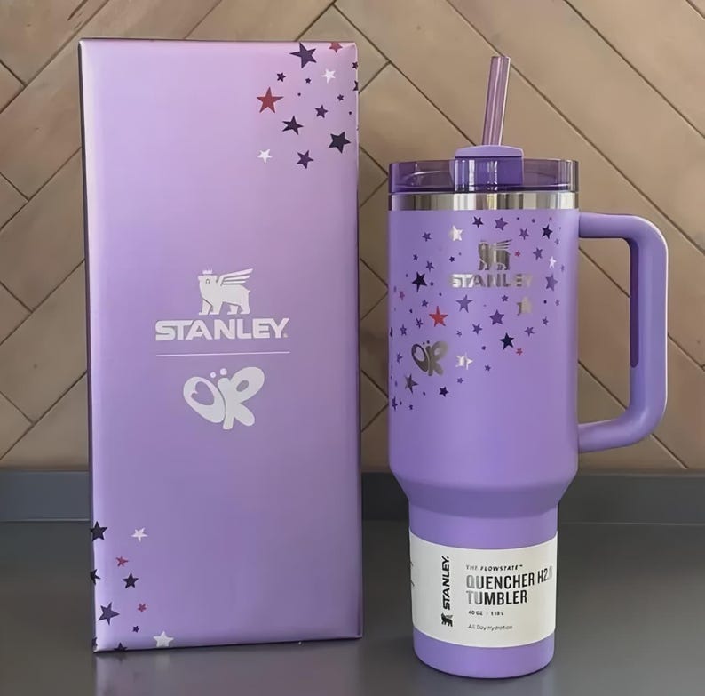 Puede incluir: Un vaso Stanley Quencher H2.0 Flowstate lavanda con asa y pajita, junto a una caja a juego. El vaso y la caja est&aacute;n decorados con gr&aacute;ficos de estrellas y mariposas. El vaso tiene una etiqueta blanca con texto negro.