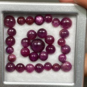 以下が含まれることがあります： さまざまなサイズの、研磨された丸いルビーの宝石のコレクション。正方形の透明なプラスチック容器に展示されています。ルビーの色は、濃い赤色から紫がかった赤色まであります。