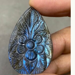 May include: A teardrop-shaped, carved gemstone with a floral design. The stone features a central flower and surrounding leaf patterns. The stone has a blue and gray iridescent color.