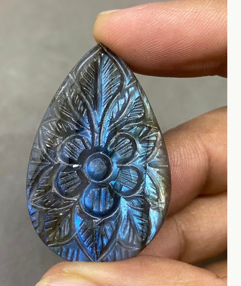May include: A teardrop-shaped, carved gemstone with a floral design. The stone features a central flower and surrounding leaf patterns. The stone has a blue and gray iridescent color.