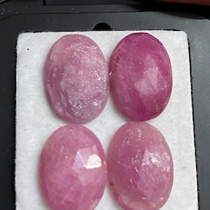 May include: Four oval-shaped, faceted gemstones in varying shades of pink and red are displayed on a white surface within a black container. The gemstones have a polished, reflective surface, showcasing their internal structure and color variations.