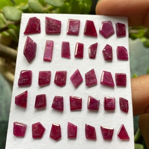 May include: A collection of thirty small, faceted, deep pink to red gemstones arranged in five rows on a white rectangular display card. Each stone features a unique, irregular geometric shape with visible surface textures and varying angles.
