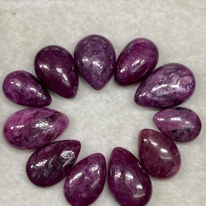 May include: Ten pear-shaped gemstones in a variety of shades of purple, arranged in a circle on a white surface.