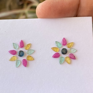 May include: Two sets of loose gemstones arranged in a circular pattern. Each set has nine gemstones in shades of pink, yellow, green, and blue. The center of each circle has a dark blue gemstone.