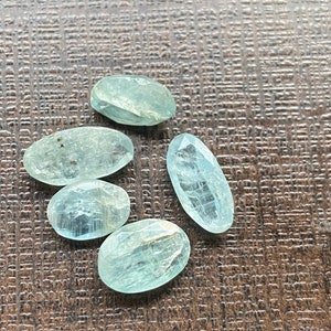 Könnte beinhalten: Fünf ovale, hellblaue Edelsteine mit facettierten Oberflächen liegen auf einer braunen, strukturierten Oberfläche. Die Edelsteine haben eine durchscheinende Qualität, fangen das Licht ein und zeigen subtile Farbvariationen. Die Anordnung deutet auf eine Sammlung loser Edelsteine hin.