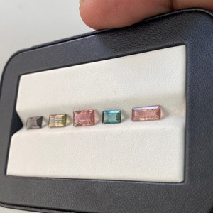 May include: Five rectangular cut gemstones in a black display case. The gemstones are arranged in a row from left to right. The gemstones are a light blue, a light green, a pink, a light brown, and a dark blue.