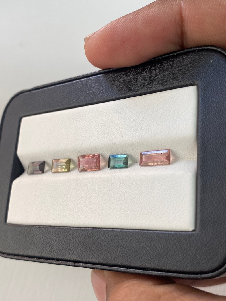 May include: Five rectangular cut gemstones in a black display case. The gemstones are arranged in a row from left to right. The gemstones are a light blue, a light green, a pink, a light brown, and a dark blue.