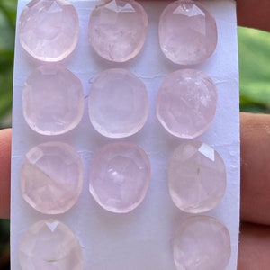 Könnte beinhalten: Elf ovale Rosenquarz-Edelsteine mit facettierten Oberflächen. Die Steine haben einen blassrosa Farbton und sind auf weißem Hintergrund angeordnet. Jeder Edelstein hat ein einzigartiges Innenmuster.