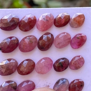 Puede incluir: Una colección de 15 piedras preciosas ovaladas facetadas de color rosa y rojo. Las piedras preciosas están dispuestas en tres filas de cinco sobre una superficie blanca.