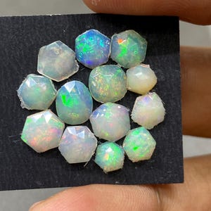 May include: A collection of faceted opals in various shapes and sizes. The opals display a range of colors, including white, blue, green, and flashes of other colors. The gemstones are arranged on a black surface.