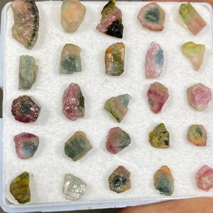 May include: A collection of 20 small, colourful, and translucent gemstones in a white box. The gemstones are various shades of pink, green, and black.