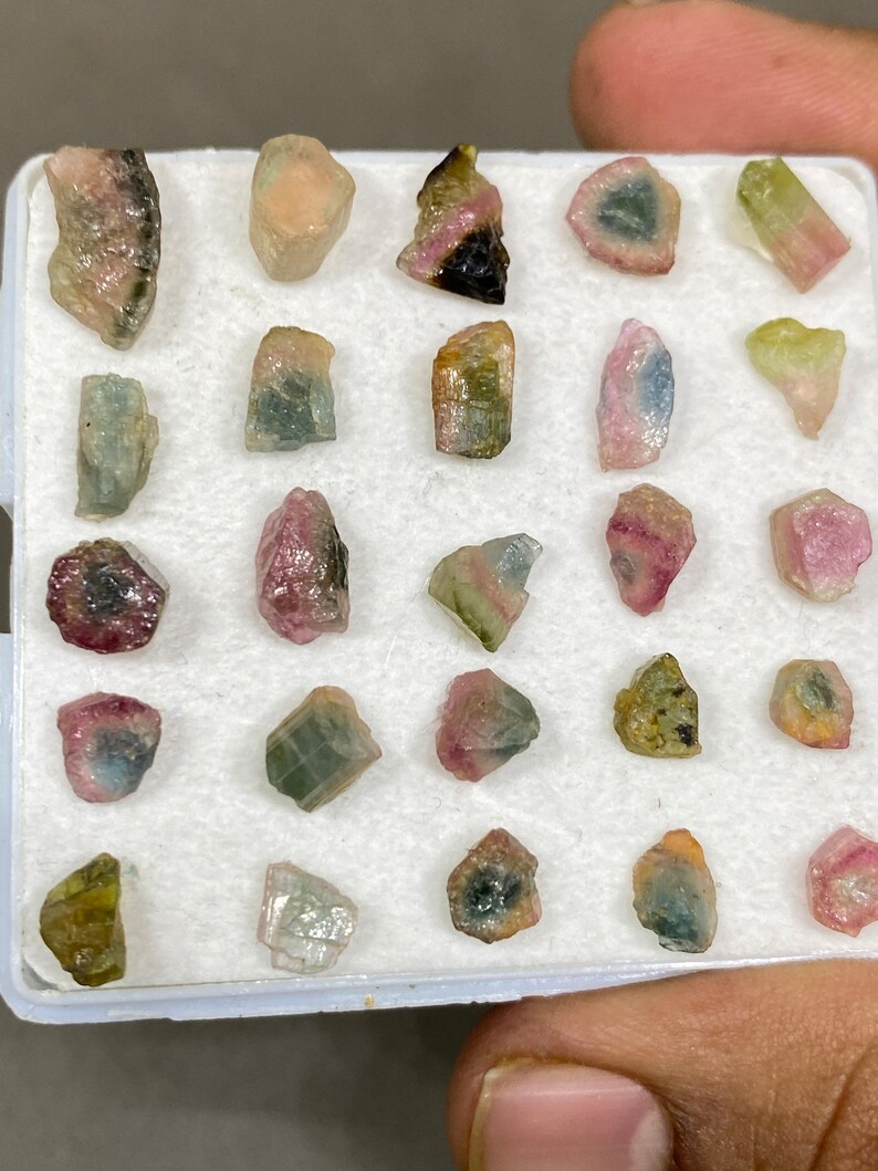 May include: A collection of 20 small, colourful, and translucent gemstones in a white box. The gemstones are various shades of pink, green, and black.