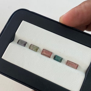 May include: A close-up of six rectangular gemstones in a black and white display case. The gemstones are various shades of green, pink, and gray.