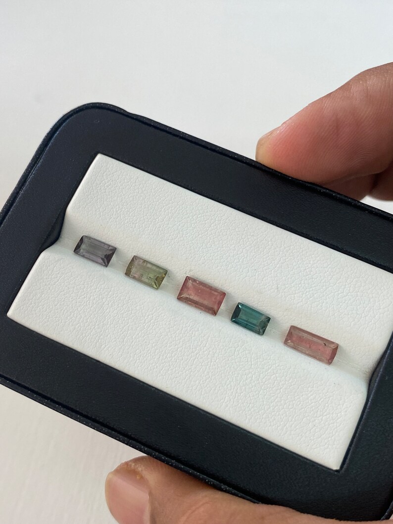 May include: A close-up of six rectangular gemstones in a black and white display case. The gemstones are various shades of green, pink, and gray.