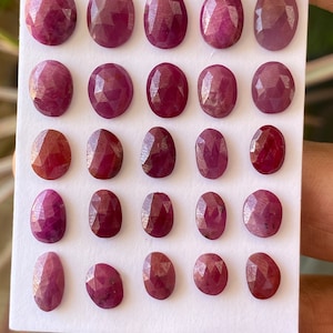 May include: A collection of twenty-one faceted oval-shaped rubies, each displaying a deep red hue. The gemstones are arranged on a white surface, showcasing their polished surfaces and internal clarity. The rubies vary slightly in size and shade.