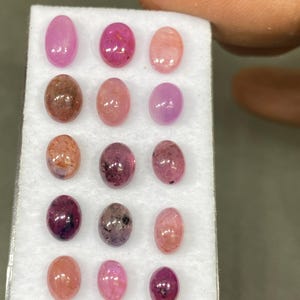 May include: A collection of oval-shaped gemstones in shades of pink, red, and purple. The gemstones are arranged on a white surface, showcasing their varying colors and translucence. The gems are held by a person's fingers.