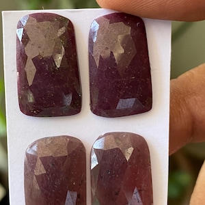 May include: Four faceted, rectangular-shaped, deep red gemstones. The gems are held on a white card, with a hand holding the card. The gems have a polished, reflective surface and are likely rubies.