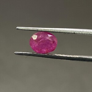 May include: A faceted, oval-shaped ruby gemstone held by tweezers. The ruby is a deep, vibrant pinkish-red color, with visible internal inclusions. The background is a neutral gray.