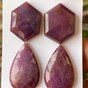 Puede incluir: Dos rubíes facetados hexagonales y dos rubíes facetados en forma de pera con un tono rojo violáceo intenso. Estas cuatro piedras preciosas sueltas presentan una superficie pulida con inclusiones naturales visibles sobre una tarjeta blanca.