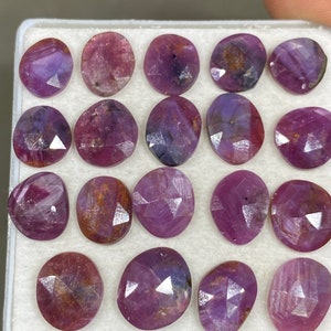 May include: A collection of 18 oval-shaped, faceted gemstones in shades of purple and pink. The gemstones are arranged in a white box.
