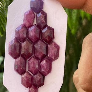 Puede incluir: Un conjunto de gemas facetadas, hexagonales, de color rojo intenso, dispuestas en un racimo sobre una superficie blanca. Las gemas varían ligeramente de color, algunas aparecen más moradas. La disposición está sujeta por los dedos de una persona.