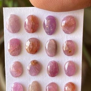 Puede incluir: Una colección de dieciséis piedras preciosas rosas pequeñas, de forma ovalada y facetadas, dispuestas en una cuadrícula de cuatro por cuatro sobre una tarjeta rectangular blanca. Cada piedra muestra tonos de rosa con inclusiones naturales.