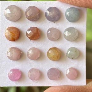 May include: A collection of faceted gemstones in various pastel colors, including pink, peach, lavender, and pale green. The gemstones are round and set on a white card, showcasing their polished surfaces and unique hues. The gems are held by a finger.