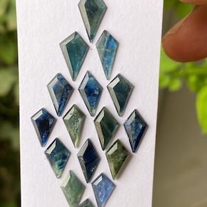 May include: A selection of kite-shaped gemstones in shades of blue and green, presented on a white card. The gemstones are faceted, showcasing diverse colour depths and clarity. The arrangement is held by a person's fingers.