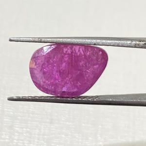May include: A vibrant pink gemstone with a faceted cut. The gemstone is held in place by a pair of tweezers.