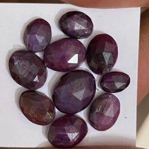 Puede incluir: Una colección de ocho piedras preciosas ovaladas y facetadas en tonos profundos de púrpura y rojo oscuro. Cada piedra presenta una superficie pulida con múltiples facetas geométricas que reflejan la luz, dispuestas en un grupo.