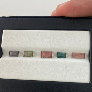 May include: Five rectangular cut gemstones in a black display case. The gemstones are arranged in a row from left to right: gray, green, pink, blue, and pink.