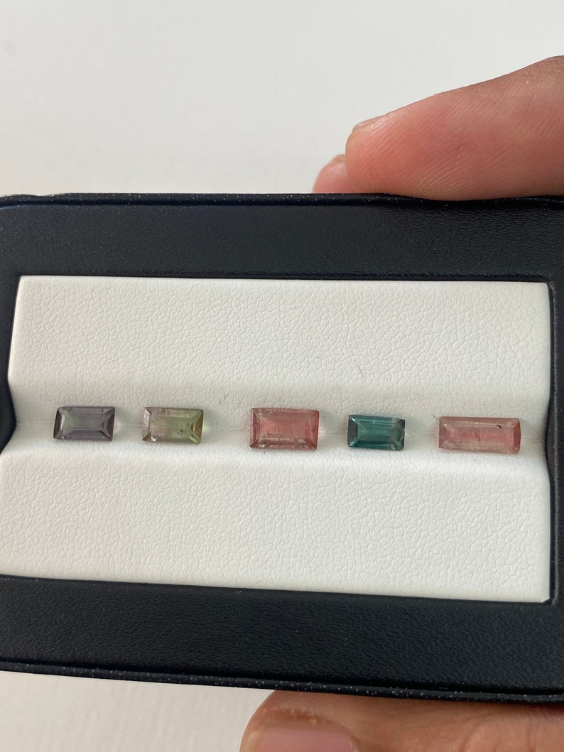 May include: Five rectangular cut gemstones in a black display case. The gemstones are arranged in a row from left to right: gray, green, pink, blue, and pink.