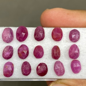 May include: A collection of 12 faceted ruby gemstones in a white display case. The rubies are a deep pink color and have a variety of shapes and sizes.