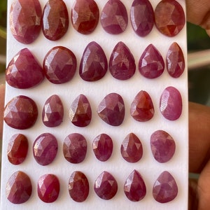 Puede incluir: Una colección de veintinueve piedras preciosas rojas facetadas y sueltas dispuestas en cinco filas sobre una tarjeta de presentación rectangular blanca. Cada piedra presenta una superficie pulida con múltiples facetas y formas variadas.