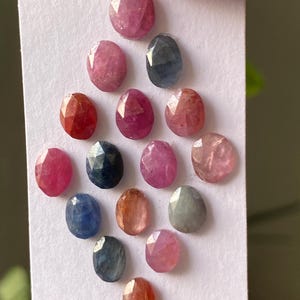 Pode incluir: Uma coleção de pedras preciosas ovais em várias cores, incluindo rosa, vermelho, azul e pêssego. As pedras preciosas são dispostas em um cartão branco, mostrando suas superfícies facetadas e variações naturais.