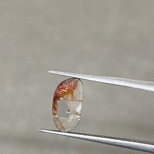May include: A small, polished gemstone held by tweezers. The stone is a translucent, light color with reddish-brown inclusions on one end. The shape is a rounded rectangle. The tweezers are silver-colored.