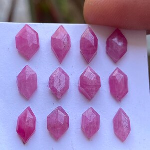 Peut inclure: Une collection de douze pierres précieuses roses facettées. Chaque gemme a une forme de diamant avec de multiples facettes, créant un effet scintillant. Les gemmes sont disposées sur une surface blanche, mettant en valeur leur couleur vibrante et leurs coupes complexes. Les gemmes sont tenues par un doigt.