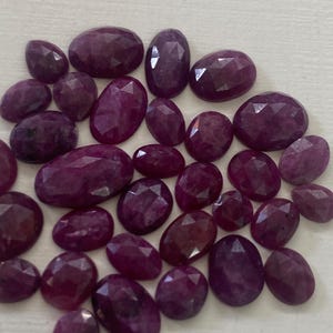 Puede incluir: Una colección de piedras preciosas de rubí sueltas, facetadas y de forma ovalada en varios tamaños. Cada piedra muestra un tono rojo magenta profundo y saturado con una superficie pulida y multifacetada que refleja la luz en cada gema.