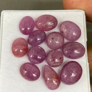Könnte beinhalten: Eine Sammlung von 10 rosafarbenen ovalen und birnenförmigen Edelsteinen. Die Edelsteine sind in einer weißen Schachtel angeordnet.