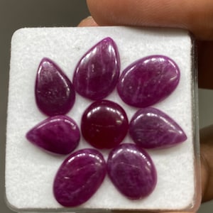Könnte beinhalten: Eine Sammlung von acht violetten Edelstein-Cabochons in verschiedenen Formen, darunter Tropfen- und Ovalformen, die auf einem weißen Schaumstoffuntergrund präsentiert werden.