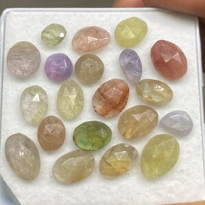 Può includere: Una collezione di 18 gemme sfaccettate in varie tonalità di verde, giallo, rosa e viola. Le gemme sono esposte in una scatola bianca.