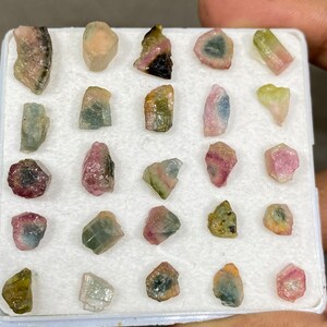 May include: A collection of 20 small, colourful, and translucent gemstones in a white foam tray. The gemstones are various shades of pink, green, and black.