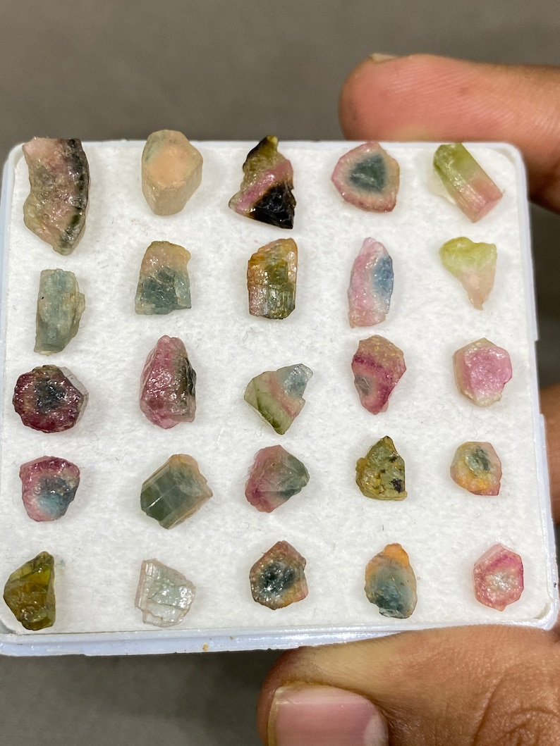 May include: A collection of 20 small, colourful, and translucent gemstones in a white foam tray. The gemstones are various shades of pink, green, and black.