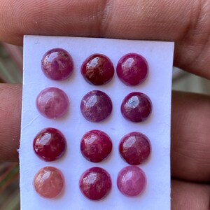 Op de afbeelding: Een verzameling van twaalf ronde, in cabochon geslepen edelstenen, gerangschikt in een raster van vier bij drie op een witte rechthoekige kaart. De stenen vertonen verschillende tinten roze, magenta en dieprood met een glad oppervlak.