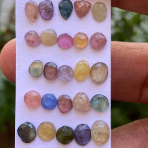 May include: A collection of 20 faceted gemstones in various colours, including pink, blue, green, and yellow, arranged in rows on a white card.