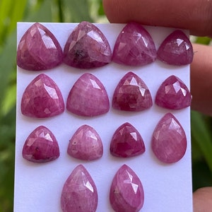 May include: A collection of 14 pink faceted gemstone cabochons in various shapes, including pear and triangle. The stones are arranged on a white card.