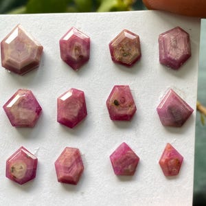 May include: A collection of faceted, pinkish-red gemstones with geometric shapes. The gemstones are arranged on a white surface, showcasing their varying sizes and internal patterns. The gems have a polished, translucent appearance.